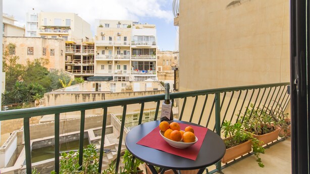 A balcony with a table holding a bowl of oranges, potted plants, and a bottle of wine. - The Strand - Apartment By The Sea Sliema (Sliema)