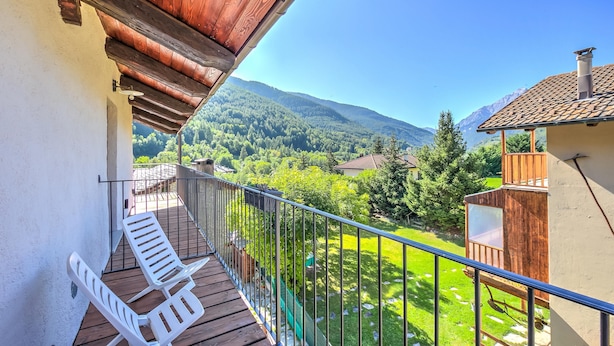 Ein Balkon mit Holzboden, weißen Stühlen und Blick auf eine grüne Landschaft mit Bergen.