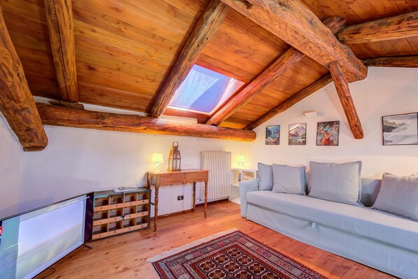 A cozy loft bedroom with a sloped ceiling, wooden beams, a skylight, a white sofa, a TV, and a wooden chest of drawers.