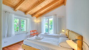Ein Schlafzimmer mit zwei Betten, Holzbalkendecke, großen Fenstern mit Vorhängen, einer Nachttischlampe und einem Heizkörper.