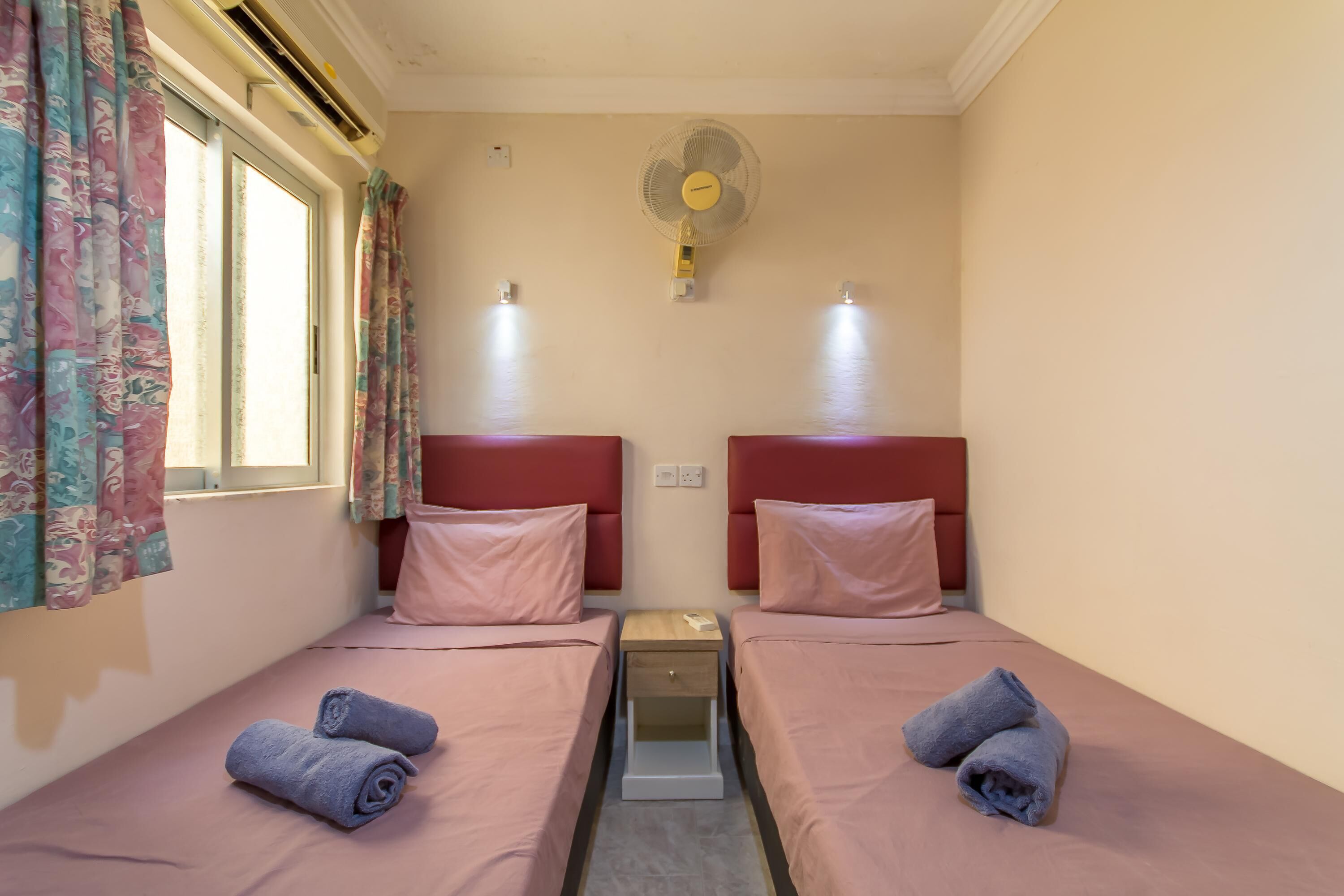 A small, cozy room with two single beds, a nightstand between them, and a wall-mounted fan.