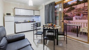Une cuisine compacte avec des meubles blancs et un four à micro-ondes, un coin repas avec une table et des chaises, et un balcon offrant une vue sur une colline.