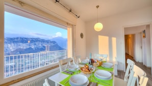 A dining area with a table set for two, a view of mountains, and a balcony with a railing. - Villa Panorama With Private Pool (Nago-Torbole)