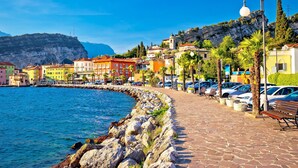 A coastal town with colorful buildings, a stone-paved promenade, and parked cars along the waterfront. - The Peace Of The Lake (Arco)