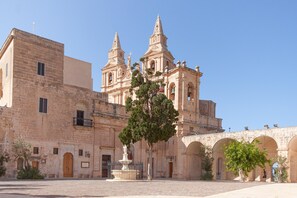 A historic building with a prominent bell tower and arches, surrounded by a paved courtyard. - Merill Pool and Sea Charming Apart 3 (Mellieha)
