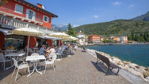 An outdoor café with tables and chairs, a stone-paved walkway, and a view of a body of water and buildings in the background. - Muri Di Arco in Arco (Arco)