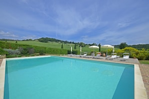 A rectangular swimming pool with a clear blue water surface, surrounded by a brick deck. There are several white lounge chairs and umbrellas arranged along the poolside. The background features a lush green landscape with rolling hills and a clear blue sky.