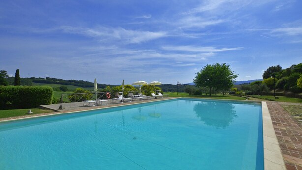 A rectangular swimming pool with a clear blue water surface, surrounded by a brick border. There are lounge chairs and umbrellas along the poolside, and a grassy area with trees and hills in the background. - Pozzovecchio in Casole d Elsa (Casole d'Elsa)