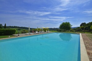 Une piscine rectangulaire à l’eau d’un bleu limpide, bordée d’une margelle en briques. Des chaises longues et des parasols sont disposés le long du bord de la piscine, et une zone herbeuse avec des arbres et des collines se trouve à l’arrière-plan.