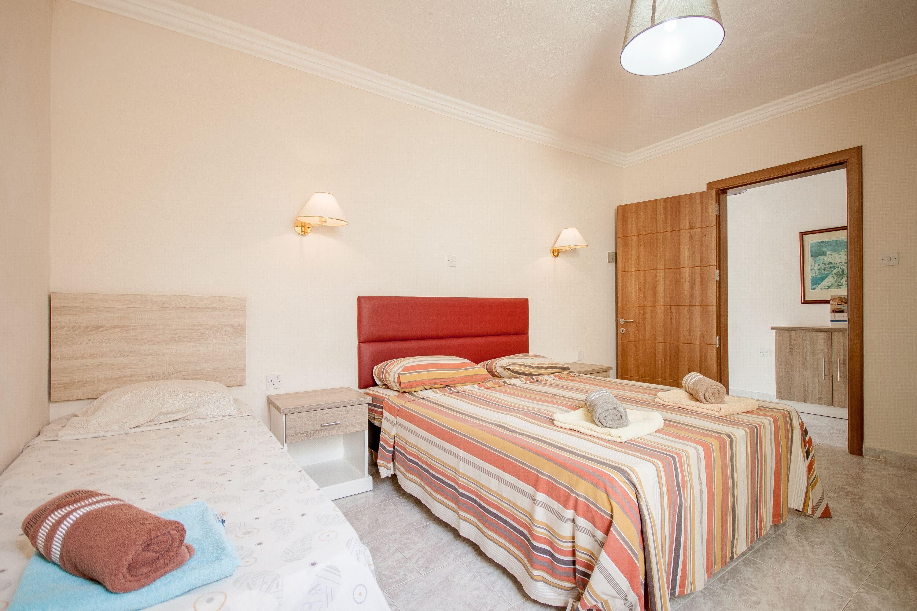 A bedroom with two beds, a wooden wardrobe, and two wall-mounted lamps.