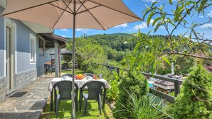 An outdoor patio with a table and chairs, a large umbrella, and a view of a lush garden and hills in the background. - Zio Enry Garden Orta Lake (Armeno)
