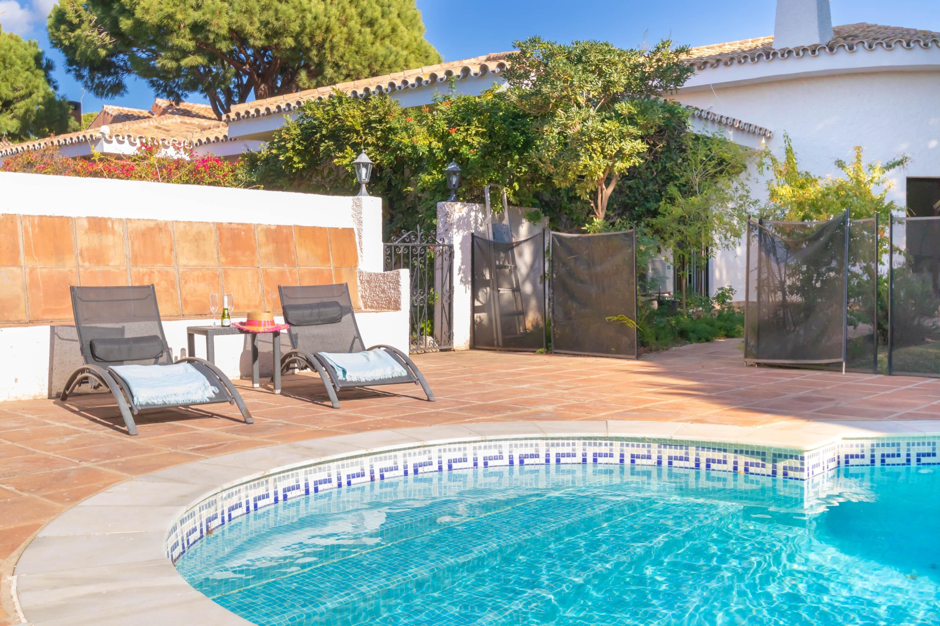 Une piscine avec deux chaises longues et une table, entourée d’un mur blanc et d’une clôture.
