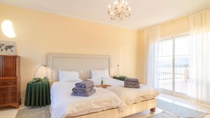A bedroom with a large bed, a chandelier, a view of the outdoors, and a green side table with a tray of towels. - Villa Carlos Mijas Golf (Mijas)
