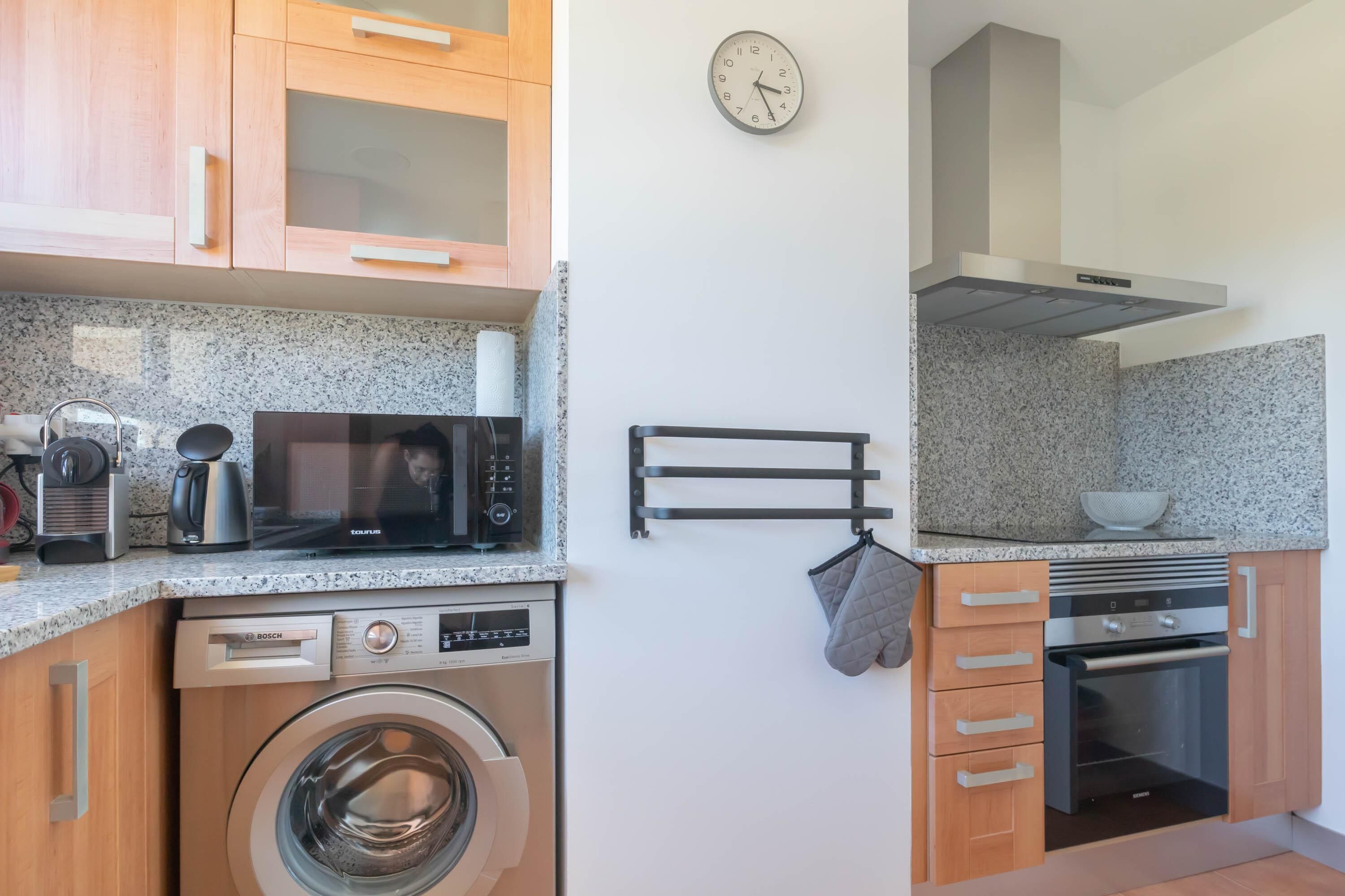 Une cuisine compacte équipée d’un lave-linge, d’un four à micro-ondes, d’un four et de meubles de rangement en bois.