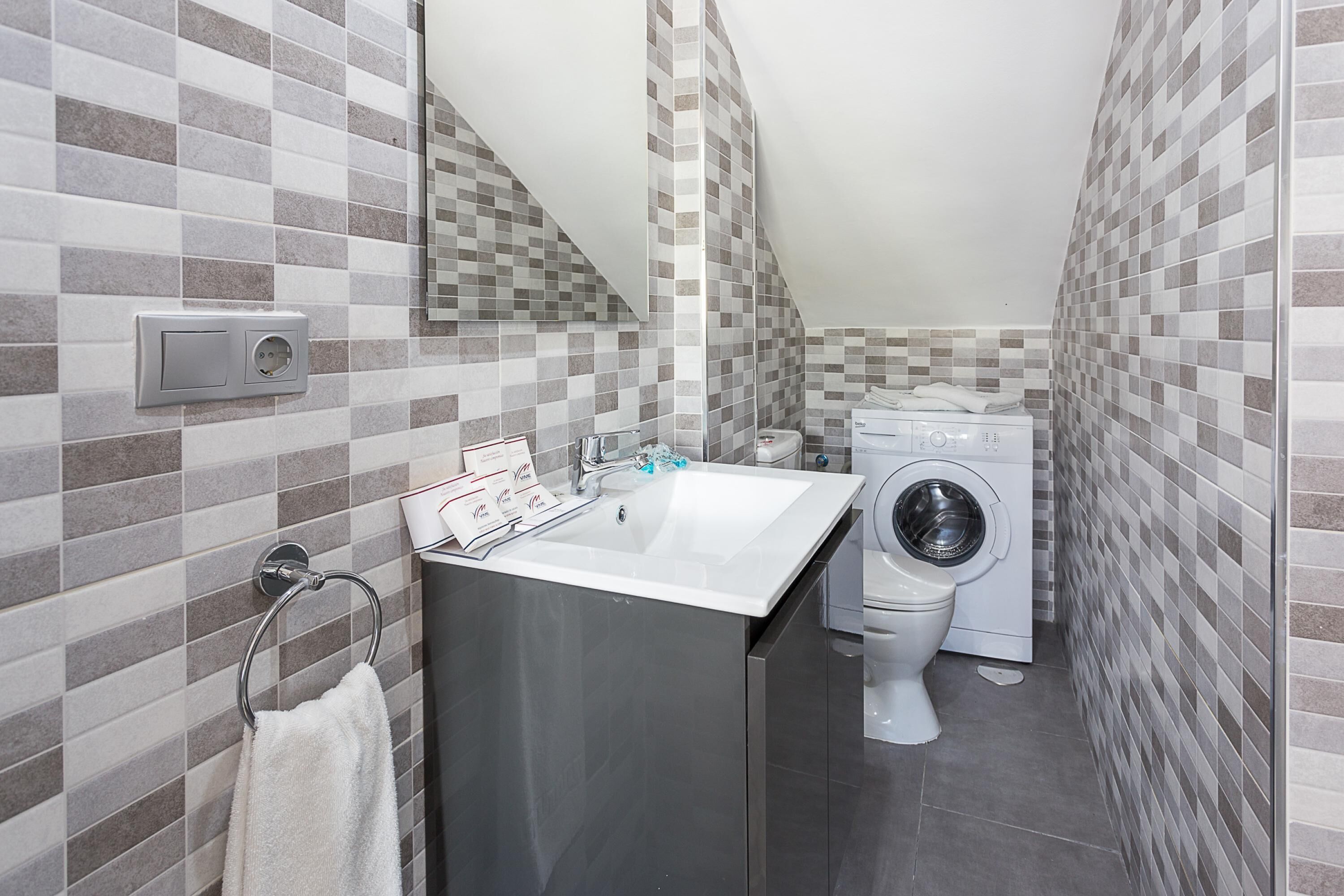 Une salle de bain moderne avec un mur recouvert de carreaux, un lavabo, un miroir, un porte-serviettes, un lave-linge et des toilettes.