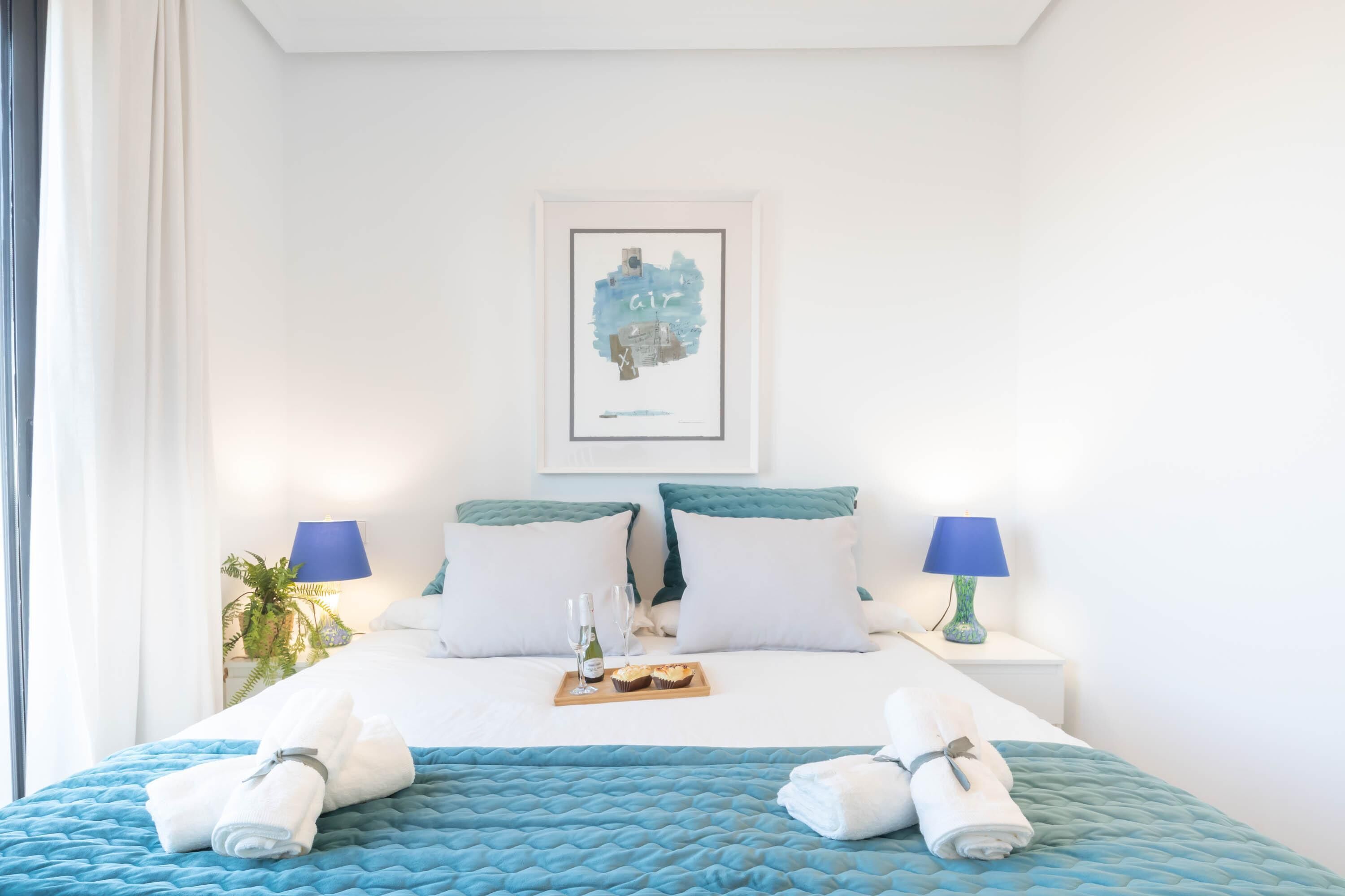 A neatly made bed with a blue quilt, two pillows, a framed artwork above, and a tray with a bottle and glasses on the nightstand.