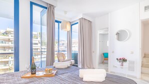 Ett modernt hotellrum med en stor säng, ett träbricka med champagne och glas, en flätad fåtölj och en balkong med utsikt över staden.