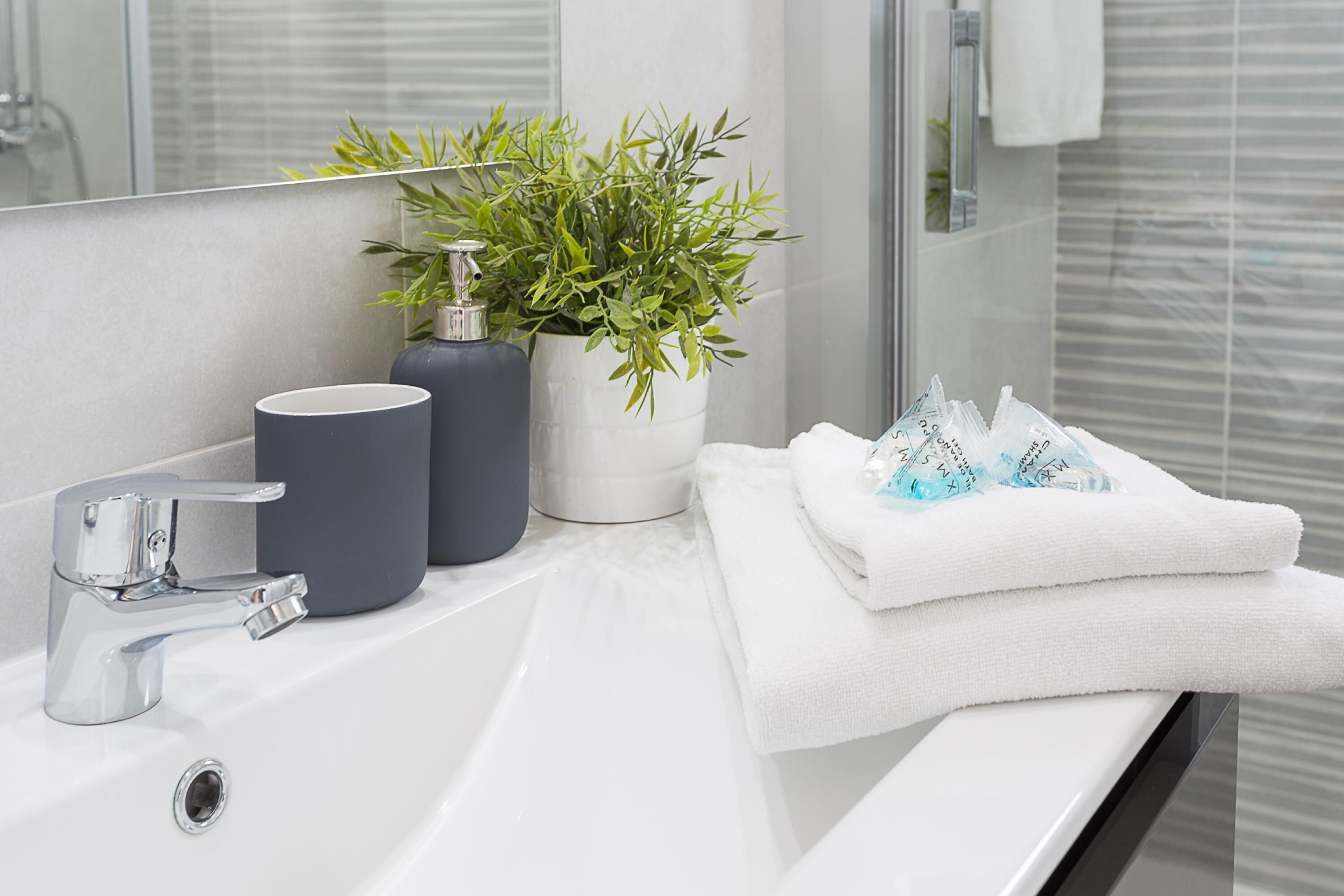 A modern bathroom with a white sink, a chrome faucet, a potted plant, and a stack of white towels.