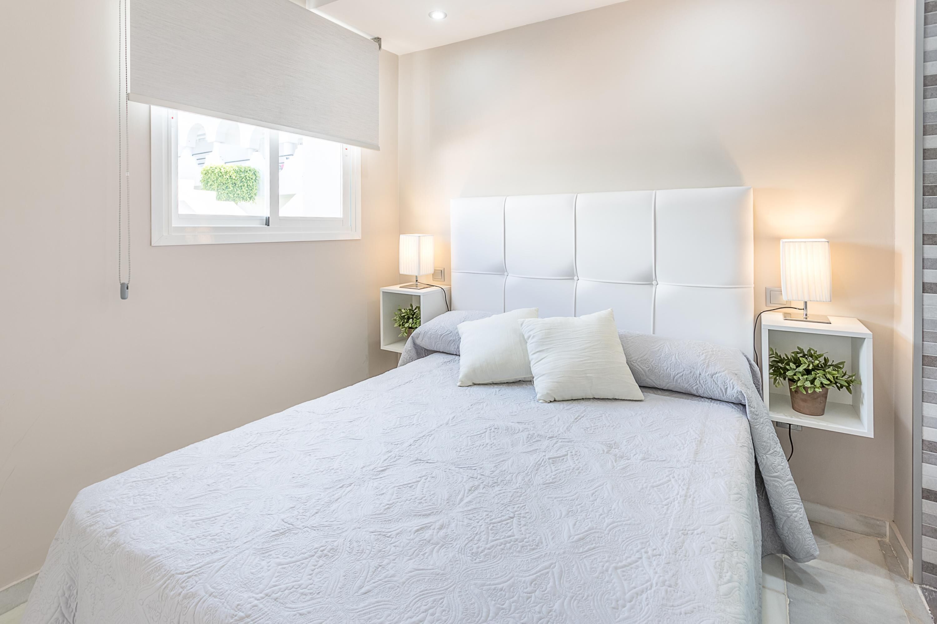 A bedroom with a large bed, two bedside tables with lamps, a window with blinds, and a potted plant on one of the tables.