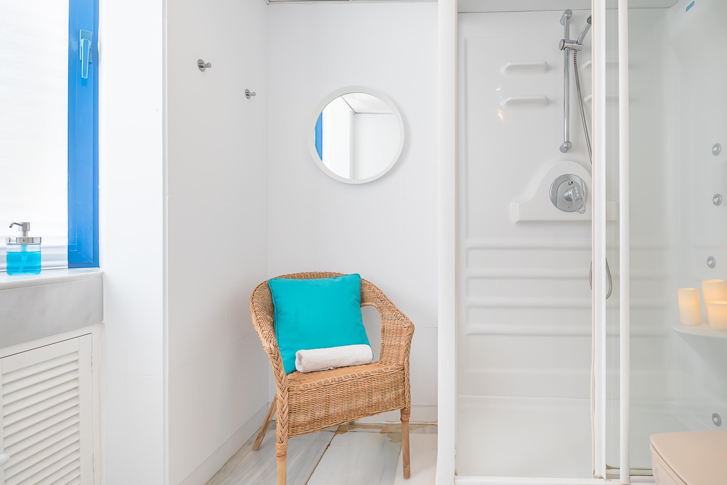 Une salle de bain avec un fauteuil en osier, un miroir rond et une douche fermée par une paroi de verre.