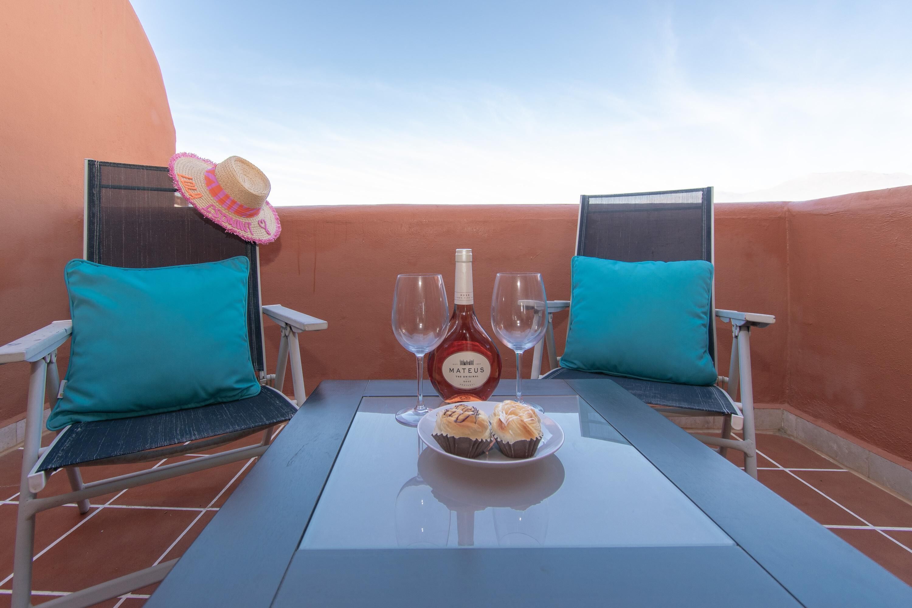 Ein Balkon mit zwei Stühlen, einem Tisch, einer Flasche Mateus Wein, zwei Gläsern und einem Teller mit zwei Backwaren.