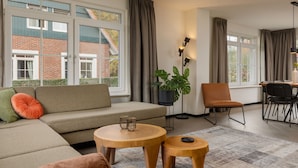 A modern living room with a sofa, two round wooden tables, a potted plant, and a dining area with a chair and table. - Home w/ Sauna & Garden (Bad Bentheim)