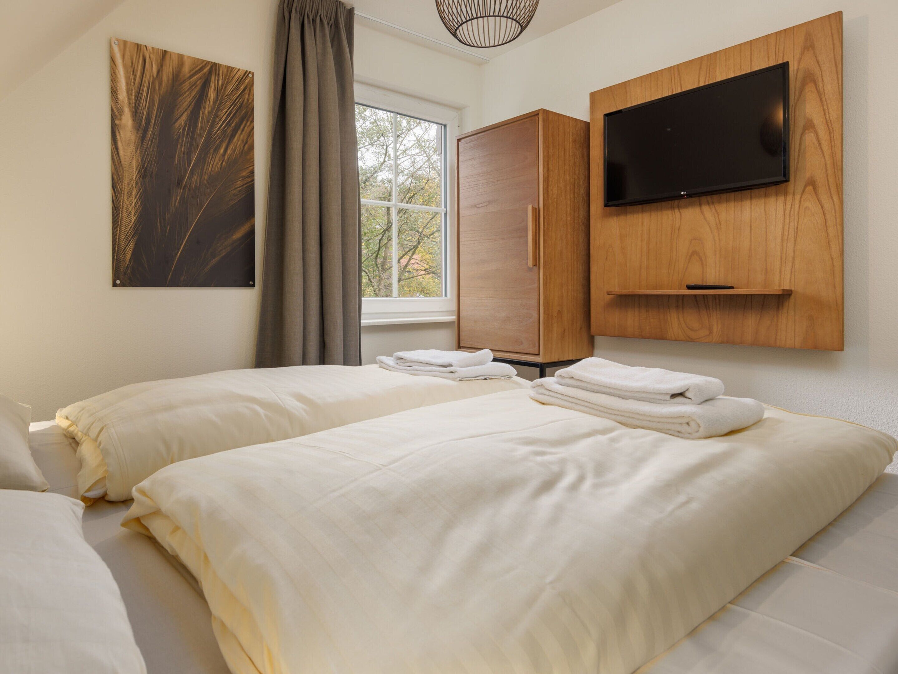 A hotel room with two beds, a flat-screen TV mounted on a wooden panel, a window with curtains, and a painting on the wall.