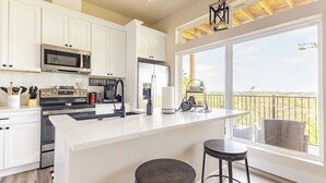 Une cuisine moderne avec des meubles blancs, un grand îlot central doté de tabourets, un coin café et une vue sur le paysage grâce à de grandes fenêtres.