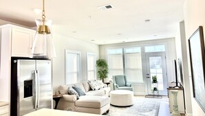 A modern kitchen with a stainless steel refrigerator, a dining area with a marble table, a beige sofa, and a large window with blinds.