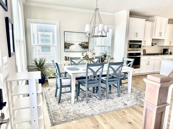 A modern kitchen with white cabinets, a dining area with a white table and blue chairs, and a large window with blinds.