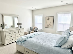 A bedroom with a bed, dresser, mirror, windows, and artwork.