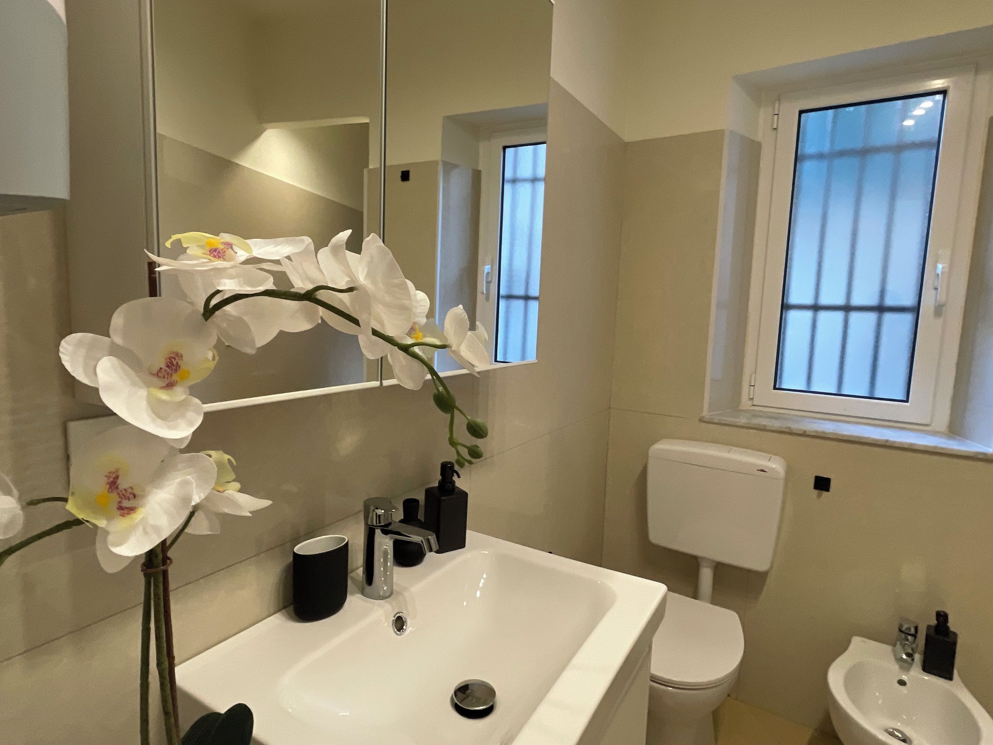 A modern bathroom with a white sink, a black cup, a mirror, and white orchids.