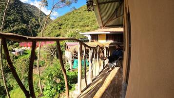 A wooden deck with a view of a mountainous landscape, a person seated on a chair, and a building with a corrugated metal roof.