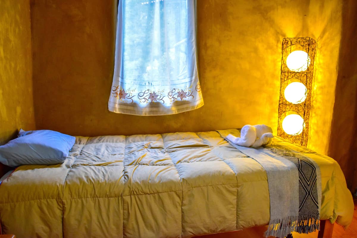 Un letto con coperta trapuntata, un cuscino e un asciugamano. Si vede una finestra con tenda e un punto luce fissato al muro.