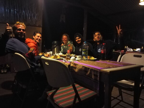 Un grupo de personas sentadas alrededor de una mesa con comida y bebidas, en un ambiente interior con poca luz.