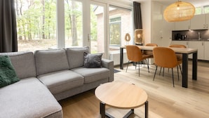 A modern living room with a gray sectional sofa, a round wooden coffee table, and a dining area with a wooden table and orange chairs. - Bungalow in De Peel by Forest Retreat (Vlierden)
