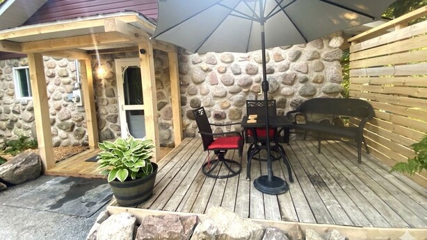 Una terraza de madera con un muro de piedra de fondo, un porche cubierto con mesa y sillas, y una planta en maceta.