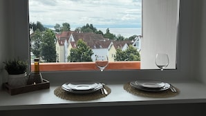 A dining area with a table set for two, a window with a view of buildings and trees, and a potted plant on the windowsill. - Apartment Emilio - Small apartment with great lake and alpine views (Überlingen)