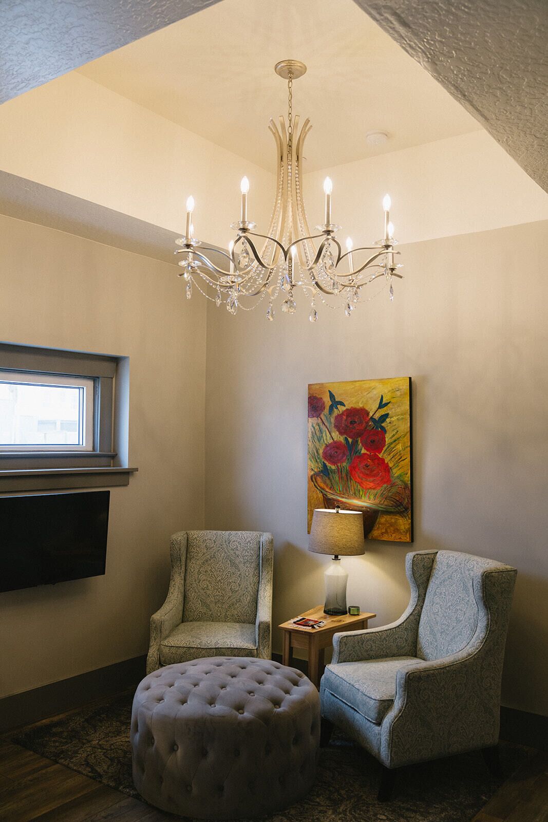 Een kamer met een kroonluchter, twee fauteuils, een rond hockertje, een schilderij en een televisietoestel.