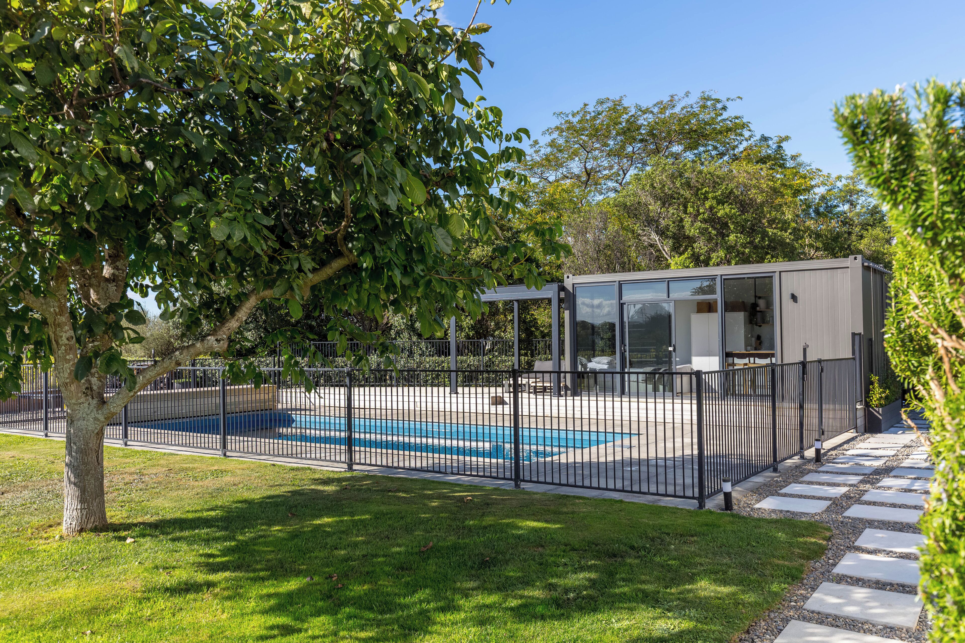 A fenced swimming pool area with a modern building, surrounded by trees and a stone pathway.