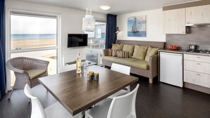 A modern kitchen with white cabinets, a dining table with white chairs, a wicker sofa, and a view of the beach through the window. - Beachfront Escape w/ Pets (Kamperland)