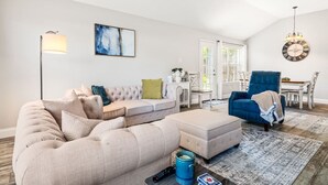 A living room with a sectional sofa, a blue armchair, a coffee table, and a floor lamp. There is a dining area with a table and chairs, and a large clock on the wall. - Extended Stay Fun Outdoor Games Gym BBQ and Dining (Lewisville)