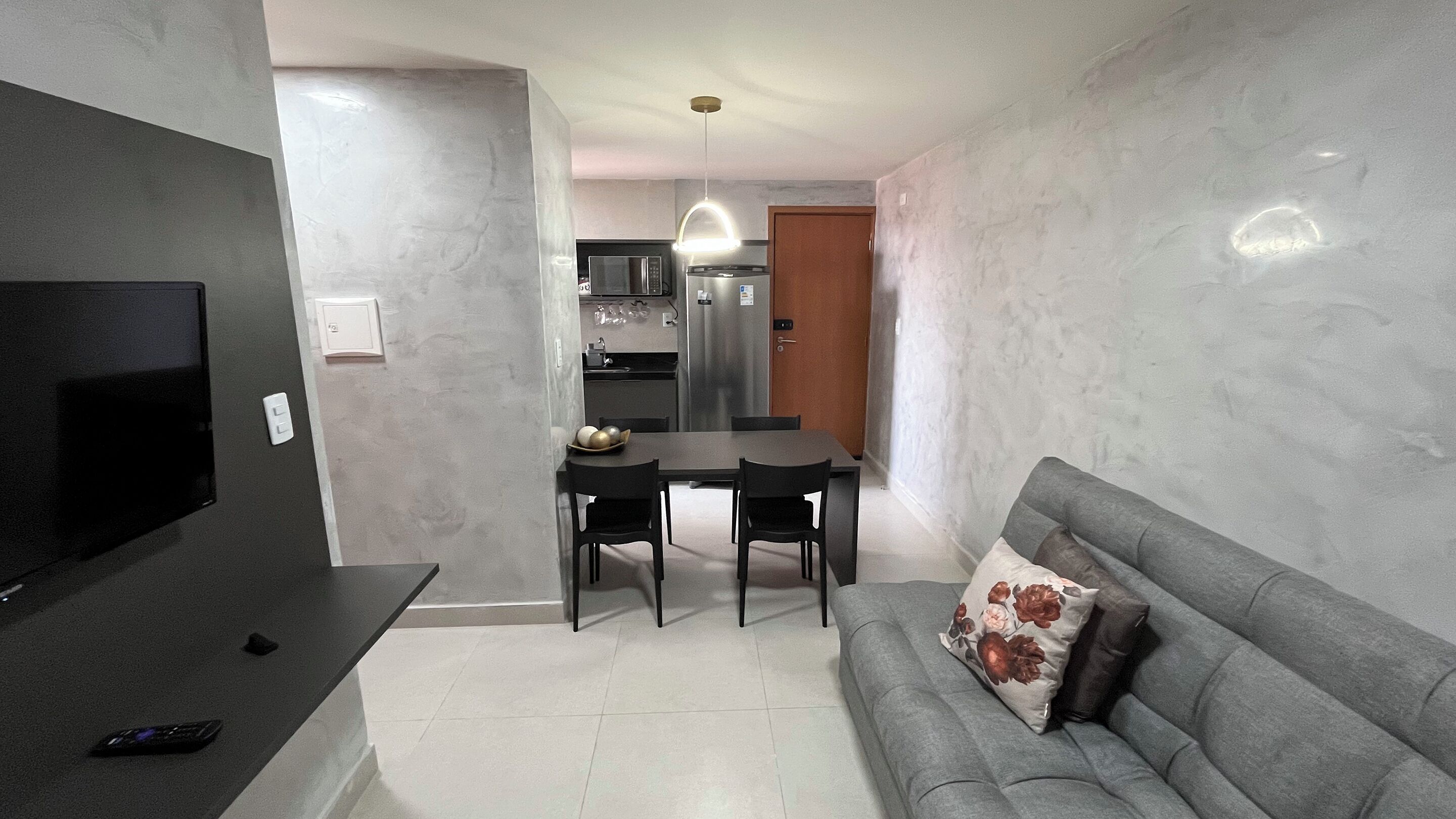 Een moderne woonkamer met een grijze bank, een flatscreen televisie en een eethoek met een tafel en stoelen.