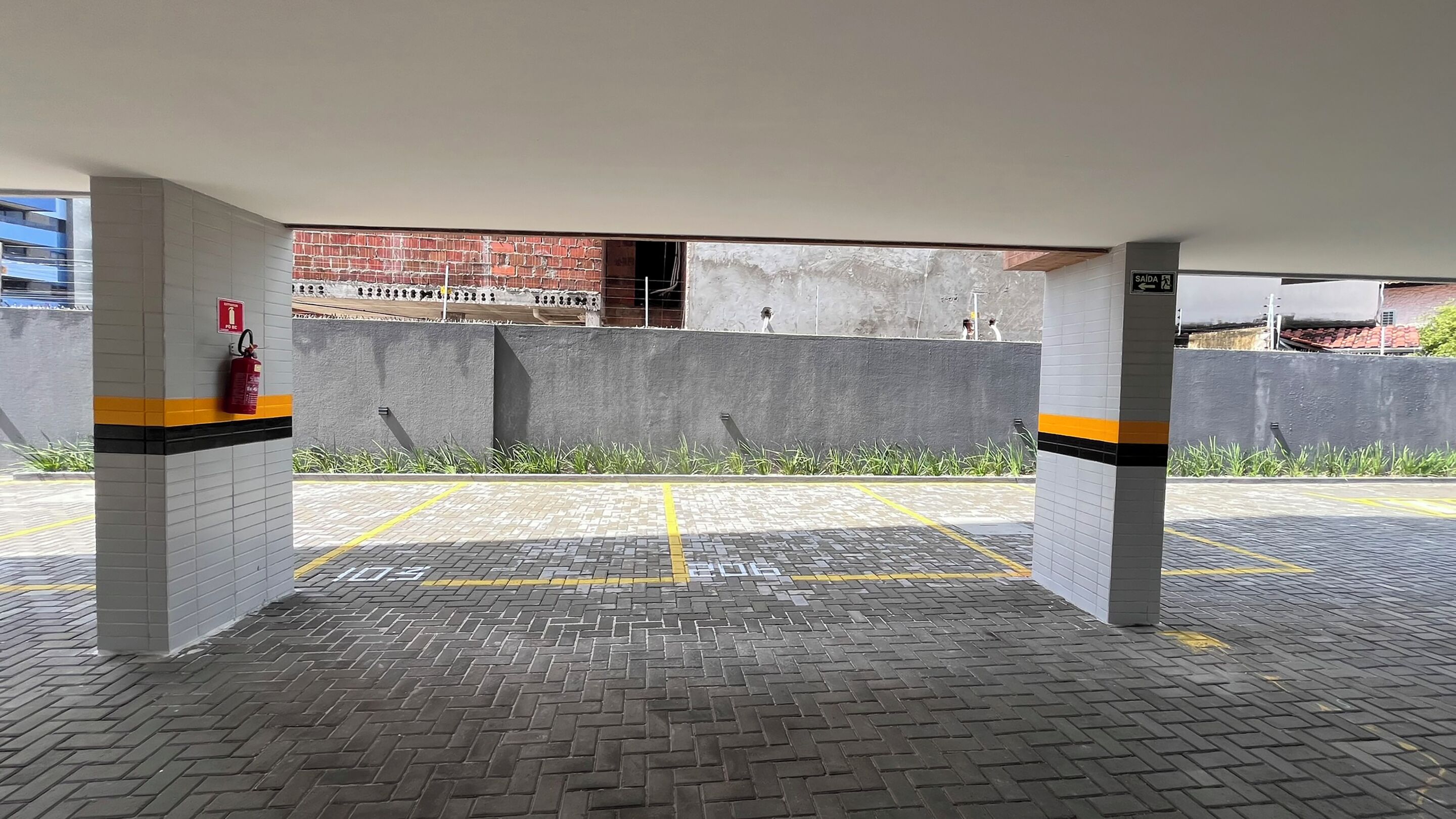 Een overdekte parkeerplaats met een brandblusser aan een pilaar, gele parkeerlijnen en een bakstenen muur op de achtergrond.