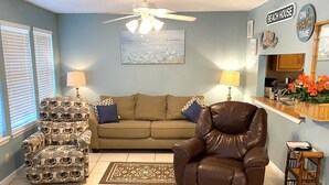 Una sala de estar con un sillón de cuero marrón, un sillón estampado, un sofá beige y una pared azul con un cuadro.