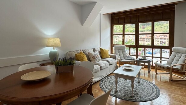 A spacious living room with a wooden dining table, a sofa with cushions, and a large window offering a view of buildings and greenery. - Apartamento Dúplex Aravedes en Potes (Potes)