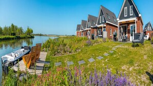 Een rij huizen met een steiger en boten, een gazon met stapelen, en een waterpartij.