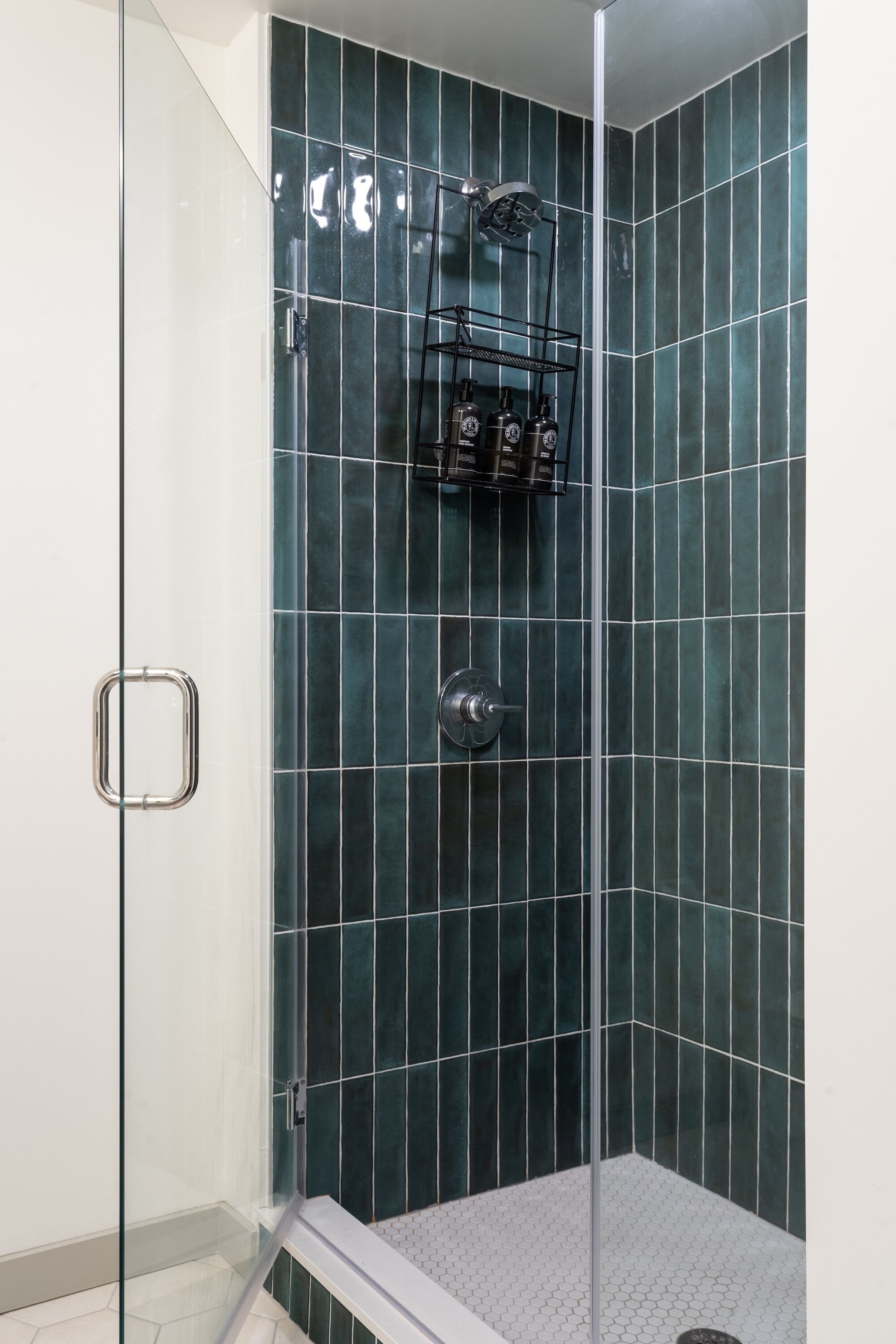 A shower with dark green tiles, a glass door, and a metal shelf with toiletries.