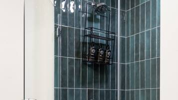 Une douche aux carreaux vert foncé, une porte en verre et une étagère en métal où sont rangés les produits de toilette.