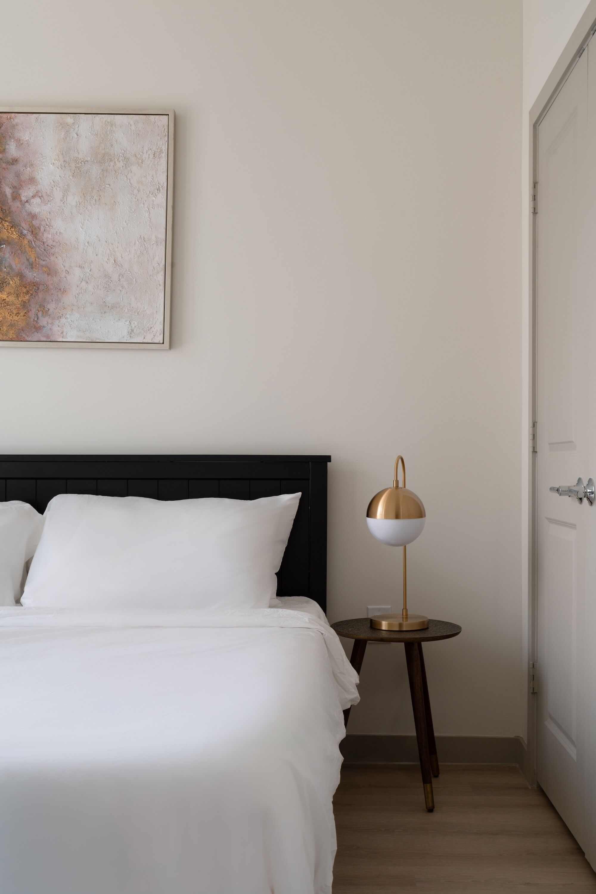 A neatly made bed with white linens, a round side table with a gold lamp, and a painting on the wall.